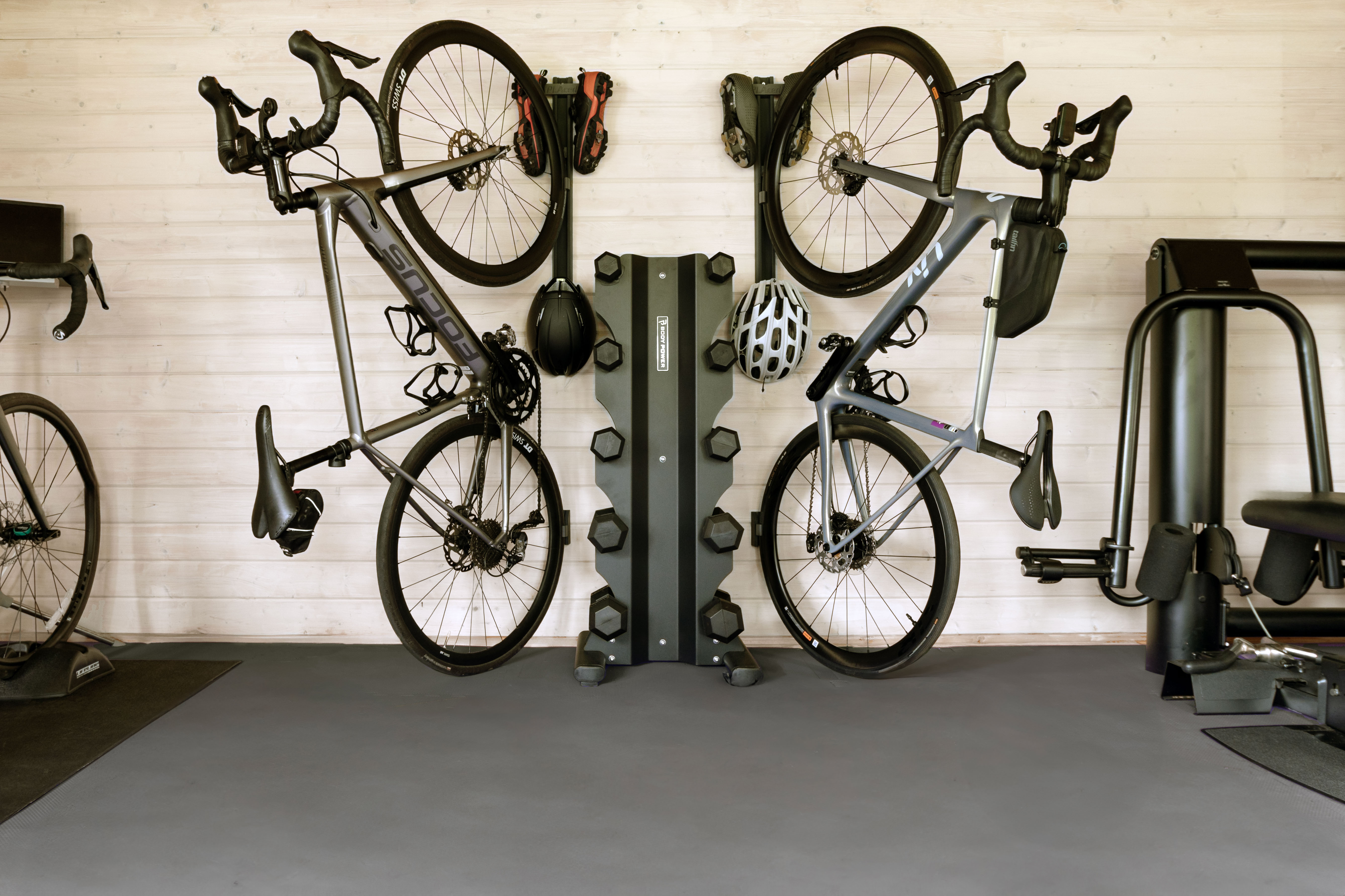 PLAced Upright Bike Storage Two road bikes mounted on the Verticale upright wall rail system in a home home fitness room, showing helmet and tyre clip accessories from PLAced