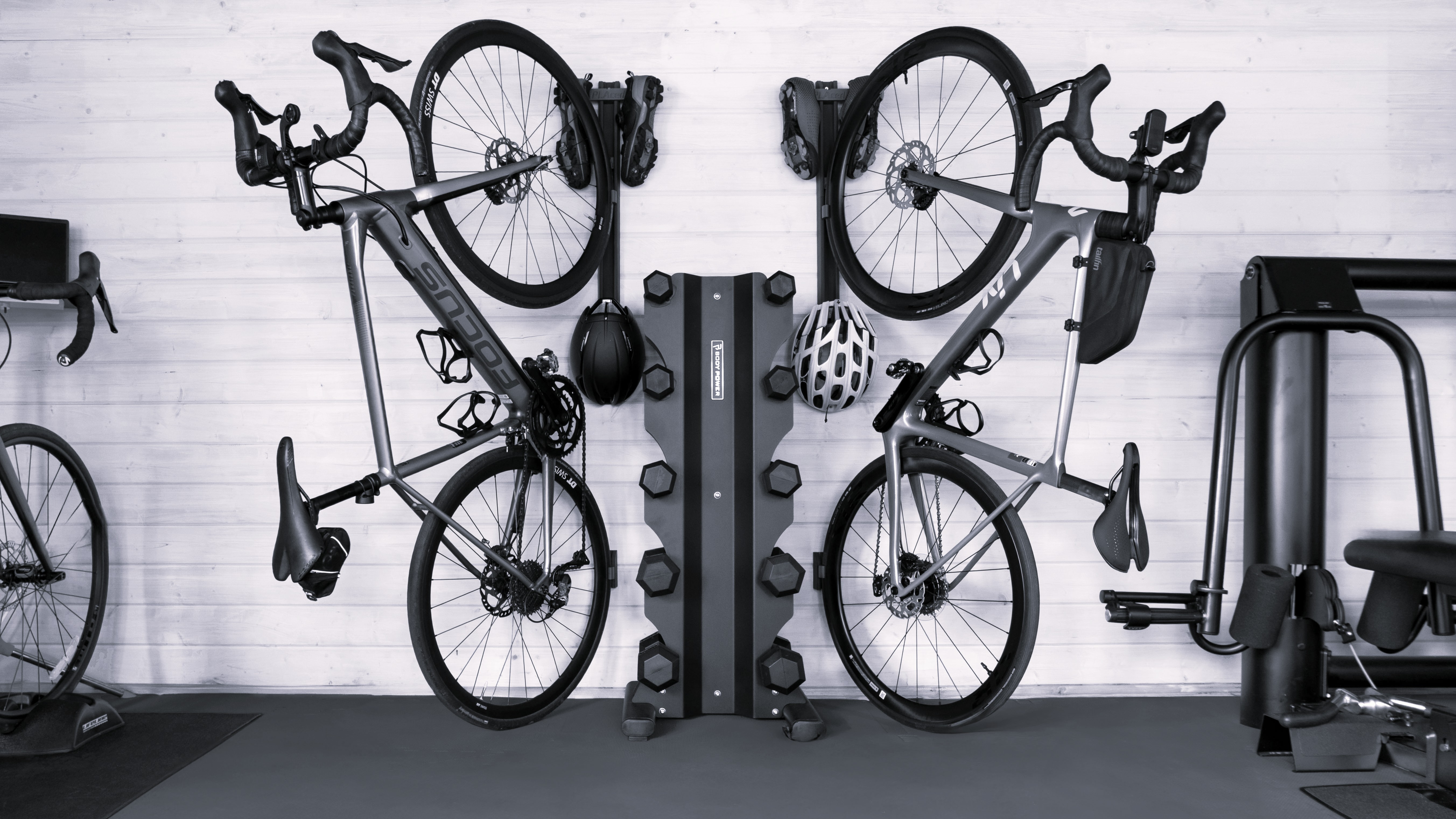 Black and white photo of Two bikes stored on the wall of a home fitness studio using the PLAced Verticale system. Between the bikes which are stored with helmet and shoes are dumbells. and gym equipment.